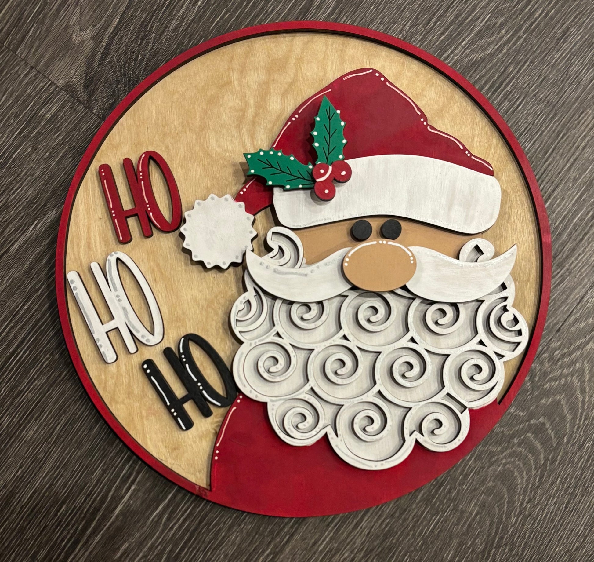 A 12" circular decorative sign featuring a stylized Santa Claus with 'HO HO HO' text around, predominantly in red and white colors.